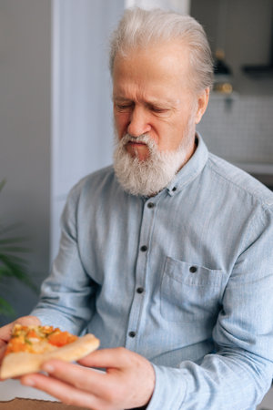 Vertical portrait of frustrated gray-haired mature adult man tasting slice of pizza with suspicion and with grimace of displeasure putting aside bad quality food, at home kitchen, close-up, cropped.の写真素材
