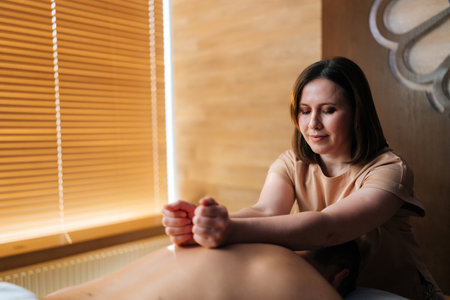 Medium shot of positive female masseuse massaging shoulder blades of muscular sportsman lying on massage table in luxury wellness center. Concept of body care and rehabilitation of spiritual peace.の写真素材