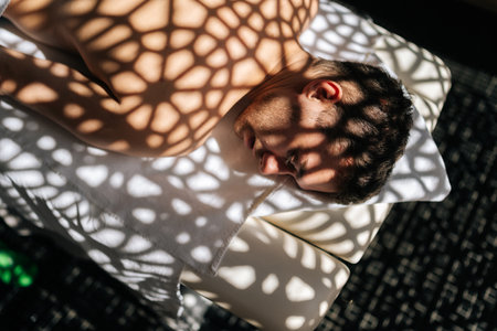 Close-up top view of handsome young man relaxing lying down with closed eyes on massage table in sunlight and beautiful shadows in spa salon. Concept of body care and rehabilitation of spiritual peaceの写真素材