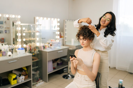 Elegance young woman client sitting in chair in beauty salon using smartphone while master hairdresser giving beauty salon service and treatment. Hairstylist doing stylish hairdo to long haired woman.の写真素材