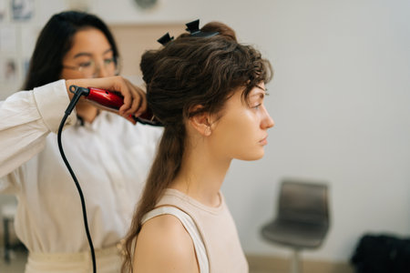 Closeup side view of professional female hairstylist using hair iron and comb for woman hairstyling in hairdressing studio. Hairdresser creating female hairdo with tongs. Concept of fashion and beautyの写真素材