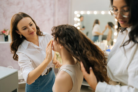 Closeup side view of smiling female makeup artist and hairstylist prepare to doing makeup and hairstyle for young woman in luxury beauty salon. Concept of four-handed makeup and styling.の写真素材