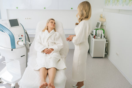 Wide shot of pretty young woman patient in white bathrobe talking to doctor during health checkup at medic clinic. Illness blonde female lying on medical couch in examination room.の写真素材