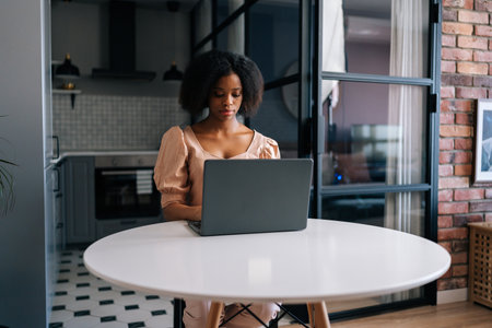 Portrait of successful African American businesswoman typing on laptop computer working in internet sitting at table in kitchen. Focused female freelancer doing online job from remote home office.の写真素材