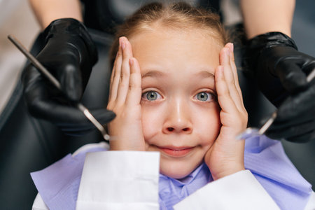 Closeup top view portrait of cute afraid child girl holding head from fear with hand, sitting on dentist chair. Close-up hands of unrecognizable dentist holding stainless steel dental tools near face.の写真素材