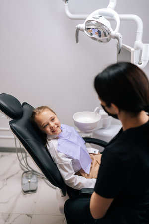 Vertical shot of unrecognizable female dentist talking with smiling child girl lying on dental chair in front of doctor, looking with happy expression. Concept of children health care treatment.の写真素材