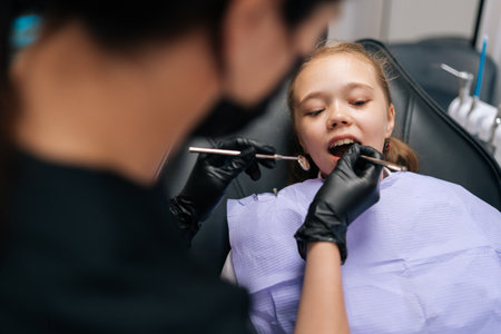 Closeup rear view of unrecognizable pediatric female dentist examining teeth of cute little girl lying on chair in dentistry clinic. Painless dental treatment, trust and professionalism of dentist.の写真素材