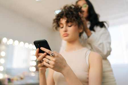 Closeup low-angle view of young woman client sitting in chair in beauty salon using phone while master hairdresser giving beauty salon service and treatment. Hairstylist doing stylish hairdo.の写真素材