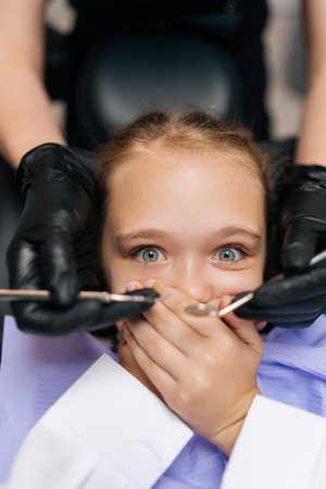 Vertical closeup portrait of afraid adorable child girl closing mouth with hands by fear in dental clinic, frightened by dental drill, afraid of treatment, looking at camera, top view , close up.の写真素材