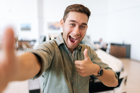 POV picture of cheerful buyer male taking selfie showing thumb up sign on smartphone from dealership after bought new car. Smiling handsome young man choosing new vehicle in showroom and making photo.の写真素材
