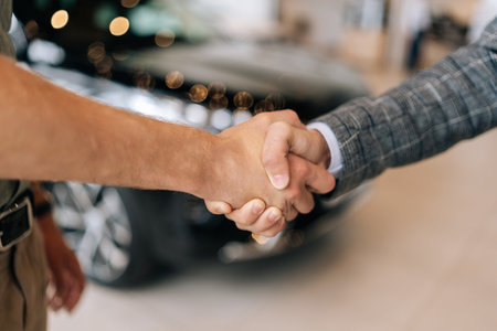 Close-up cropped shot of unrecognizable car salesman in business suit and happy male customer shaking hands purchase in auto dealership on background of auto. Concept of choosing new car at showroom.の写真素材