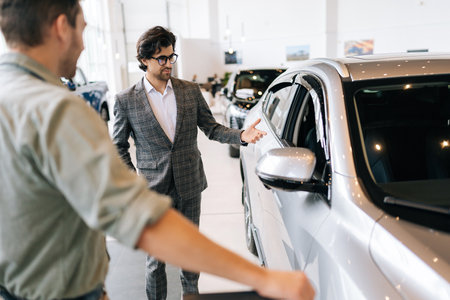 Portrait of car dealer in business suit telling unrecognizable pensive male buyer about luxurious car in dealership office. Rear view of young man client looking at auto and listening to dealer.の写真素材