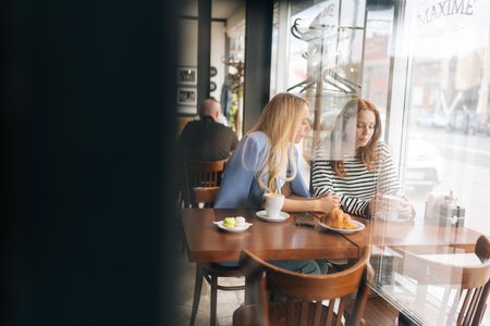 View from door glass to upset young woman telling best friend about boyfriend betrayal. Compassion showed to real best girlfriends. Life problem concept. Gossiping along with friend at coffee shop.の写真素材