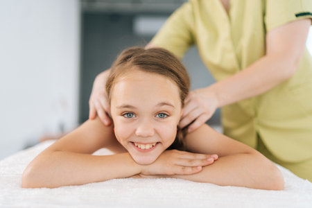 Closeup portrait of cheerful little girl having neck, shoulder and back massage by unrecognizable female pediatric masseuse at medical clinic, lying on massage table, looking at camera.の写真素材