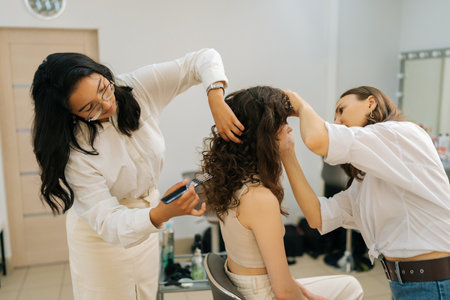 Side view of curly young woman being given applying eyeshadow in beauty salon. Hairdresser combing long hair of young woman with hairbrush. Concept of four-handed makeup and styling.の写真素材