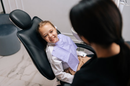 Close-up back view unrecognizable female dentist talking with lovely child girl lying on dental chair in front of doctor, looking with happy expression. Concept of children health care treatment.の写真素材