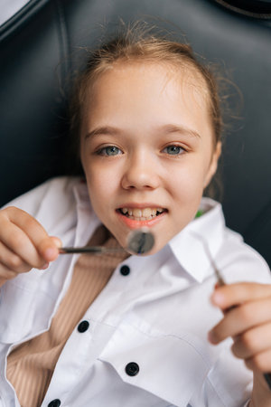 Vertical portrait of pretty little child girl holding dentistry mirror and tool lying on dental chair in stomatologist clinic, looking at camera. Concept of children teeth treatment, pediatric checkupの写真素材