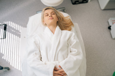 Close-up high-angle view of serene blonde female in white bathrobe lying with closed eyes on medical couch after spa treatment. Close up portrait of young woman relaxing enjoying daily hygiene routineの写真素材