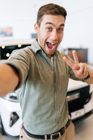 Vertical POV picture of cheerful buyer male taking selfie with victory sign on smartphone from auto dealership after bought new car. Smiling young man choosing new vehicle in showroom and making photoの写真素材
