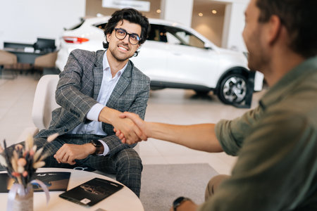 Rear view of happy unrecognizable customer male purchasing automobile in dealership signing paper shaking hands with dealer in suit enjoying successful agreement. Concept of buying auto at showroom.の写真素材