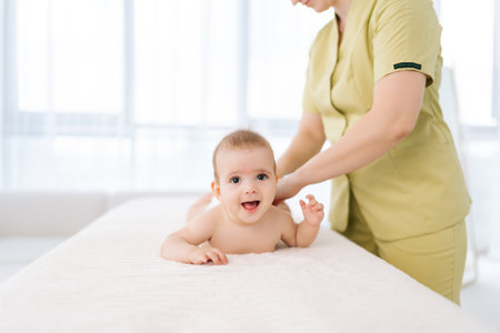 Portrait of cheerful infant baby boy lying on belly while unrecognizable female masseuse performing professional massage for back, looking at camera. Mom hands doing back massage to newborn son.の写真素材