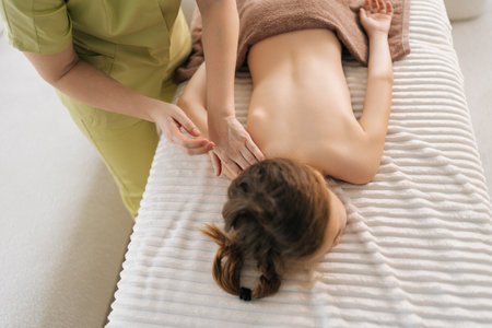 High-angle view of unrecognizable female pediatric masseuse doing therapeutic massage to relax shoulder joint and shoulder blade of little girl. Kid client get service massage in spa salon.の写真素材