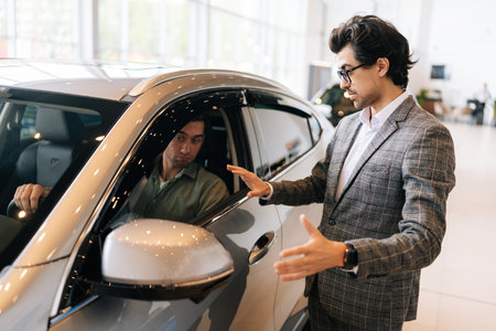 Portrait of successful customer male having consultation from to dealer representative in motor showroom sitting inside brand new car holding steering wheel. Concept of buying new auto in dealership.の写真素材