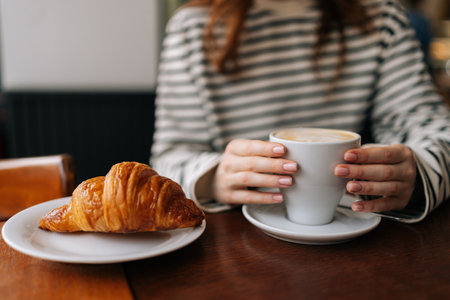 Close-up cropped shot of unrecognizable young woman holding cup of coffee sitting at table with croissant during delicious breakfast in cafe. Female hands with cup of coffee and croissant in cafeteriaの写真素材