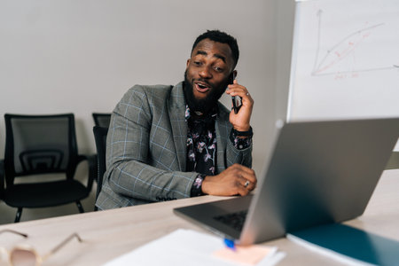 Cheerful bearded African businessman in suit talking on smartphone sitting at office desk with laptop. Happy confident black male manager consulting client about online project making business call.の写真素材
