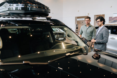 View from hood of vehicle to professional car dealer in business suit telling pensive male buyer...