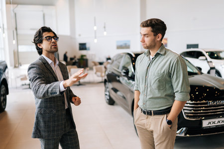 Medium shot of competent male car dealer in business suit having conversation with serious young man client at showroom. Doubtful customer guy listening manager while buying car at dealership centerの写真素材