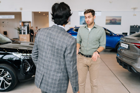 Rear view of unrecognizable professional male car seller with serious male customer in auto dealership, talking about specifications and performances of vehicle. Customers looking at cars in showroom.の写真素材