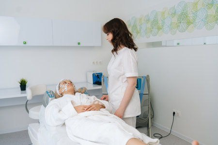 Remote view of friendly female physiotherapist communicating with lying young woman client during facial myostimulation procedure in modern beauty clinic. Concept of non-surgery cosmetology.の写真素材