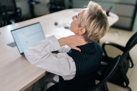 Closeup view from shoulder of tired middle-aged business woman touching, massaging neck, turning head with painful face, suffering from ache, pain, spasms in muscles after overwork at laptop on officeの写真素材