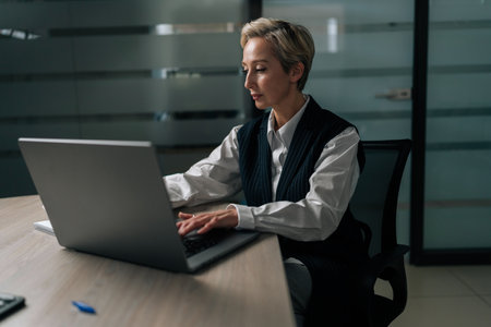 Serious elegance middle-aged 50s businesswoman sitting at modern office desk working on laptop do freelance online job. Senior adult business woman work on project typing on computer in dark room.の写真素材
