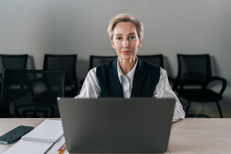 Front view of elegance middle-aged businesswoman executive top manager sitting at desk working with documents typing on laptop computer, looking at camera with serious expression, in corporate office.の写真素材