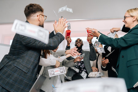 Happy excited African-American male boss in sunglasses and suit using money gun shooting with banknotes. Cheerful business team of mixed-aged diverse coworkers having fun, jumping, rejoicing victory.の写真素材