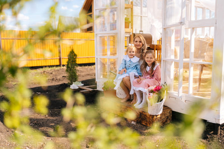 Remote view of elegance blonde young mother in dress sitting on doorstep of summer gazebo house with two little adorable daughters on sunny day, smiling looking at camera with happy expression.の写真素材