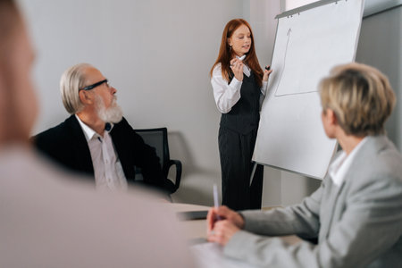 Professional female coach gives corporate presentation for businesspeople in office. Red-haired businesswoman presents new business plan on whiteboard, speaking to clients listening speaker at meetingの写真素材