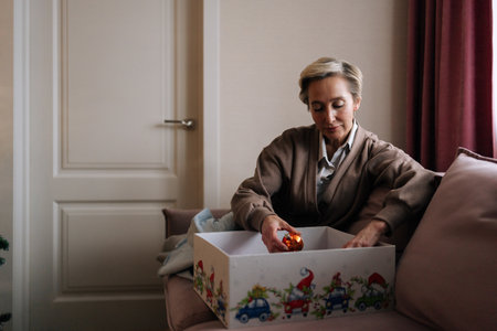 Portrait of elegant middle-aged woman choosing festive xmas tree balls in box sitting on couch. Preparation for holidays, decorates Christmas tree for New Year. Concept of home festive atmosphere.の写真素材