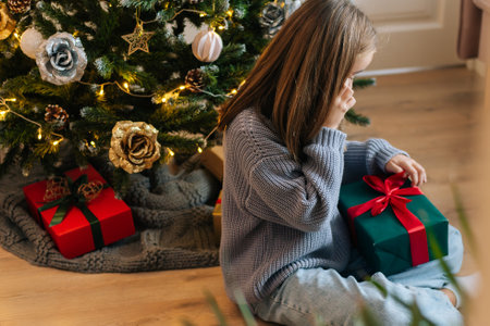 Top view of adorable little girl talking on smartphone in living room, holding and looking at wrapped gift box, calling relatives saying thanks, sitting on background of Christmas tree.の写真素材