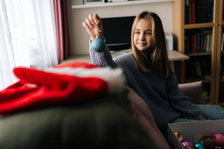 Portrait of attractive little girl preparing for holidays, choosing festive Christmas tree toys from box, decorates xmas tree for New Year in living room. Concept of home festive atmosphere.の写真素材