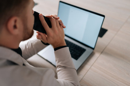 View from shoulder of serious pensive businessman in suit holding smartphone sitting at office desk with laptop computer, thinking over work project. Business owner pondering on company future vision.の写真素材