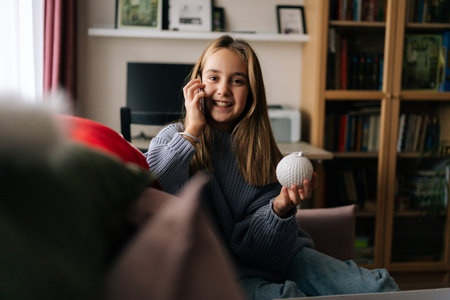 Portrait of cheerful little girl talking on smartphone, holding in hand silver festive Christmas ball. Pretty kid preparing for holidays, decorates xmas tree for New Year at home, by window.の写真素材