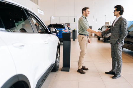 Remote side view of confident car salesman in business suit and cheerful male customer in auto dealership. Two happy people shaking hands seal purchase of new car in dealership office.の写真素材