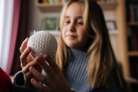 Close-up portrait of charming little girl holding and looking festive Christmas ball with happy expression. Smiling kid decorates xmas tree for New Year at home in dark living room alone.の写真素材