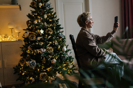 Side view of elegant middle-aged woman holding smartphone make video call on Christmas Eve congratulates relatives enjoy remote talk, standing on background of illuminated xmas tree in dark room.の写真素材