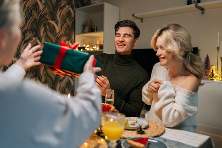 Rear view of unrecognizable mother-in-law giving festive gift box with Christmas present to happy man sitting with family at dinner feast table at home on xmas eve. Concept of home festive atmosphere.の写真素材
