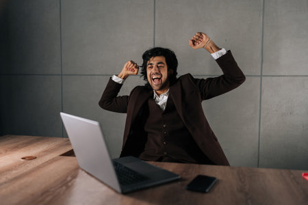 Portrait of cheerful excited businessman in suit celebrating business success achievement working at desk with laptop. Cheerful CEO feeling motivated by good work, get job, receive great news.の写真素材