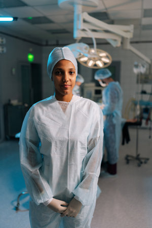 Vertical portrait of African-American female doctor in surgical uniform standing posing looking at camera in dark operating theater. Multiethnic team of surgeons performing operation on background.の写真素材